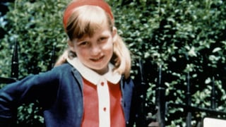 Diana als Mädchen in einem roten Kleid vor einem Zaun stehend. Sie lächelt.