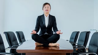 Eine Frau im Anzug sieht auf einem Sitzungtisch und meditiert.