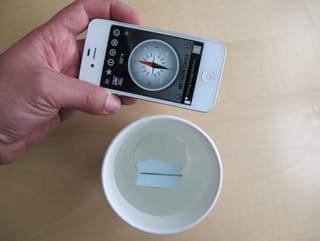 Eine Nadel schwimmt auf einem kleinen Zettelchen in einem Becher mit Wasser. Dazu hält eine Hand ein Handy, auf dem ein Kompass zu sehen ist. Nadel und Kompass zeigen in dieselbe Richtung.