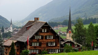 Holzhaus vor zwei Kirchen, im Hintergrund das Tal.