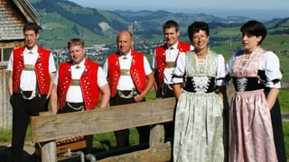 Vier Jodler und zwei Jodlerinnen in Appenzeller Trachten hinter und vor einem Holzzaun.