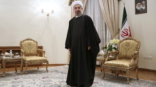 Ruhani in einem Raum mit goldenen Stühlen. Er trägt ein langes schwarzes Gewand.