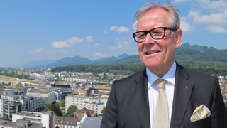 Ernst Zingg trägt einen Aanzug mit schwarzem Blazer, weissem Hemd, beiger Krawatte und beigem «Poschettli». Im Hintergrund die Martinskirche und die Stadt Olten, sowie die Jurakette.
