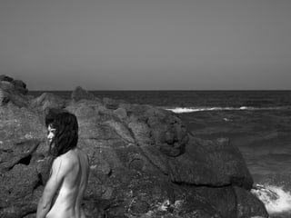 Meer mit Felsenküste, nackte Frau mit dem Rücken zum Betrachter schaut nach links.
