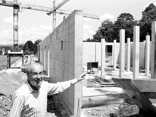 Ein Mann in gestreiftem Hemd zeigt lächelnd auf einen unfertigen Rohbau aus Beton, im Hintergrund Kräne.