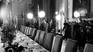 Jacqueline Kennedy im Weissen Haus an gedeckter Tafel, im Hintergrund Fotografen, Kameras, Filmlicht.