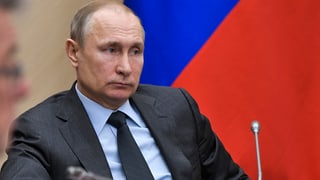 Wladimir Putin sitzt vor einer russischen Flagge, vor ihm ein Mikrofon.