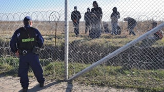 Ungarischer Bemater an Grenzzaun – dahinter sind Flüchtlinge zu sehen.