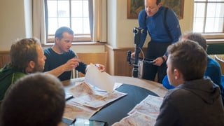 Eine Gruppe von Menschen sitzt mit Landeskarten und Wetterinformationen an einem Tisch. Hinter dem Tisch steht ein Mann mit einer Kamera. 