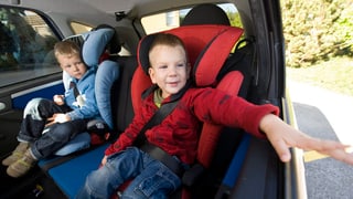 Zwei kleine Buben auf dem Rücksitz eines Autos, angeschnallt in einem roten und einem blauen Kindersitz.