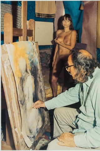 Ein Mann mit schütterem, grauen, lockigen Haar, Brille und Schnauz malt einen Akt. Im Hintergrund eine junge, nackte Frau, die ihre rechte Brust anhebt. 