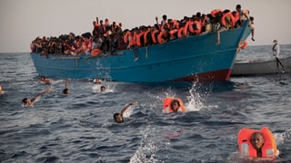 Überfülltes, blaues Boot, etliche Menschen befinden sich mit Schwimmwesten im Wasser.