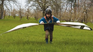 Ein kleiner Junge mit Helm rennt mit einer Flugkonstruktion über eine Wiese. 