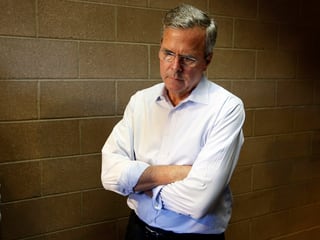 Bush steht vor einer braunen Backsteinmauer in einer nachdenklichen Pose. Er hat die Arme verschränkt und schaut nach unten.