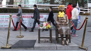 Eine Bronzestatue eines Mädchen sitzt auf einem Stuhl. Dahinter laufen Männer vorbei. 