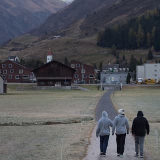 Drei Personen gehen auf einer Strasse. Im Hintergrund ist Andermatt und die Berge zu sehen.
