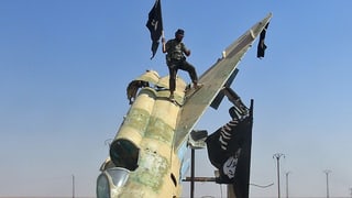Ein Mann mit einem eurpäisch anmutenden Hut hisst eine IS-Fahne auf einem eroberten Flugzeug.