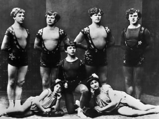 Auf einer alten Aufnahme stehen mehrere junge Männer in Kostümen in einer Reihe.