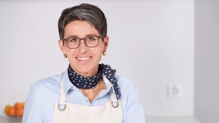Eine Frau in weisser Kochschürze mit Brille und dunkelblauem Foulard.