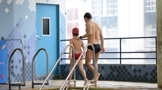 Ein junger Mann in Badehose geht am Schwimmbecken entlang, seine Hand berührt einen Jungen in Badehose am nackten Rücken. 