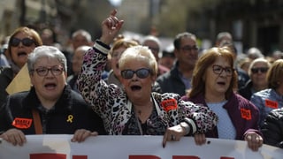 Drei ältere Frauen - zwei mit kurzen grauen Haaren, eine mit längeren braunen - an einer Demonstration. Sie halten ein Transparent und skandieren Parolen.  