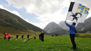 Ein Fahnenschwinger mit der Flagge des Kantons Graubünden und Alphornbläser auf einer Wiese in den Bergen.