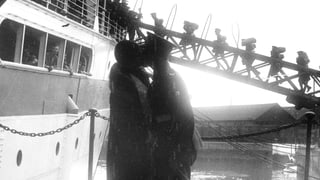 Ein Pärchen küsst sich. Er trägt Uniform. Im Hintergrund: Ein Schiff und Soldaten, die das Schiff besteigen.