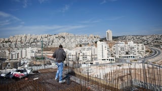 Baustelle mit Armierungseisen auf einem Hausdach, im Hintergrund eine jüdische Siedlung