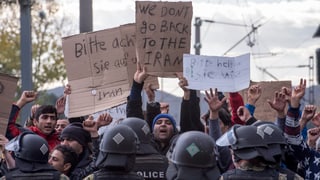Flüchtlinge halten Schilder mit Protestbotschaften hoch, davor Polizeikräfte