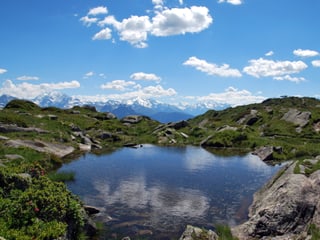 Klarer Bergsee umgeben von Wiesen und Felsen. Blauer Himmel mit wenigen Schönwetterwolken. 