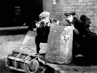 Cagney und ein Mann verstecken sich hinter einer Mülltonne und zielen mit Pistolen. Cagney und ein Mann verstecken sich hinter einer Mülltonne und zielen mit Pistolen.