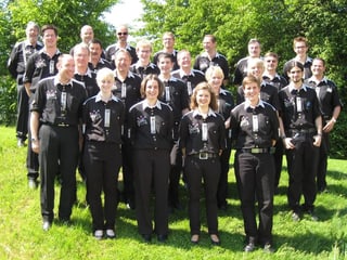 Die Blasmusikanten und -musikantinnen tragen schwarze Hemden und Hosen und posieren fürs Gruppenbild auf einer grünen Wiese.