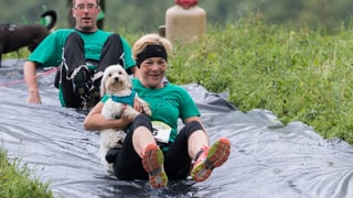 Eine Hundehalterin hält ihren kleinen Hund fest und rutscht auf einer Plastikplane den Hang herunter. Eine Hundehalterin hält ihren kleinen Hund fest und rutscht auf einer Plastikplane den Hang herunter.