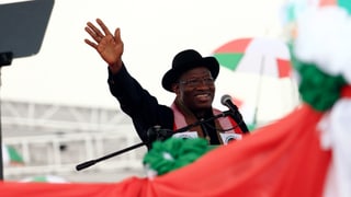 Jonathan Goodluck steht an einem Mikrofon und Winkt, er trägt schwarze Kleidung und einen Hut Jonathan Goodluck steht an einem Mikrofon und Winkt, er trägt schwarze Kleidung und einen Hut