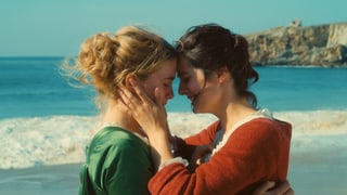 Zwei Frauen stehen nahe beieiander umarmen sich und halten ihre Köpfe nahe beieinander. Im Hintergrund sieht man das Meer.