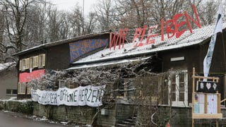 Ein zweistöckiges Haus mit dunkelbraunen Schindeln und einem rechteckigen, einstöckigen Anbau. Auf und vor dem Haus diverse Transparente. Ein zweistöckiges Haus mit dunkelbraunen Schindeln und einem rechteckigen, einstöckigen Anbau. Auf und vor dem Haus diverse Transparente.