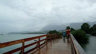 Regenwolken hängen über dem See. Auf dem Steg Leute mit Regenschirm.