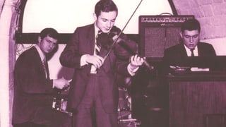 Schwarzweiss Foto. Drei Musiker spielen auf einer winzig kleinen Bühne. Vorne der Musiker Jean-Luc Ponty mit der Geige, hinten der Schlagzeugspieler Daniel Humair und daneben Eddy Louis an der Orgel.