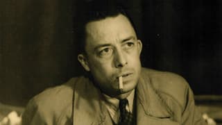 Albert Camus in einer Porträtaufnahme. Im Mantel an einem Tisch sitzend, mit Zigarette im Mund.