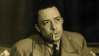 Albert Camus in einer Porträtaufnahme. Im Mantel an einem Tisch sitzend, mit Zigarette im Mund. Albert Camus in einer Porträtaufnahme. Im Mantel an einem Tisch sitzend, mit Zigarette im Mund.