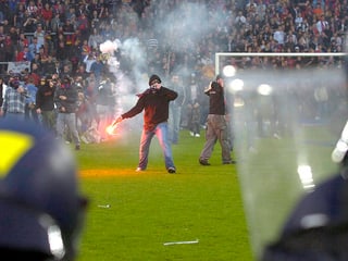 EIn Vermummter hält eine Petarde in der Hand, im Vorderung das Schutzschild eines Polizisten, im Hintergrund vermummte FCB-Fans.