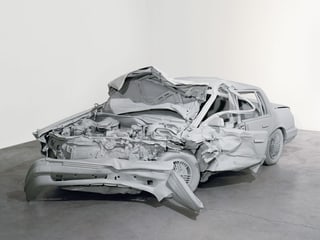 Eine hellgraue Skulptur zeigt ein Auto, dessen vorderer Teil komplett zerstört ist. 