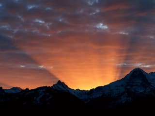 Unten Konturen von Schneebergen, der Himmel ist bewölkt und die Sonne scheint hinter der Bergkette. Helle Strahlen brechen über die Krete. 