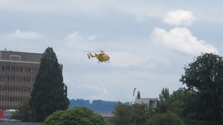 Ein  gelber Helikopter vom TCS setzt zur Landung auf das Kantonsspital Aarau an. Im Hintergrund Wolken, im Vordergrund Bäume.