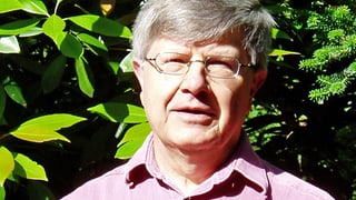 Ein Mann mit Brille und grauen Haaren, im Hintergrund ist ein Gebüsch erkennbar.