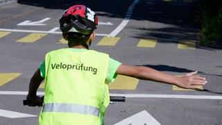 Kind mit einer West mit der Aufschrift Veloprüfung streckt den rechten Arm hinaus zum Abbiegen Kind mit einer West mit der Aufschrift Veloprüfung streckt den rechten Arm hinaus zum Abbiegen