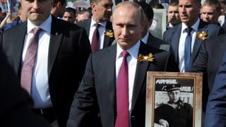 Putin in einem Strassenumzug zwischen anderen Männern, er hält ein Schwarz-weiss-Foto eines Soldaten in der Hand, es ist sein Vater im Zweiten Weltkrieg.