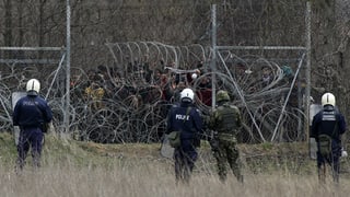 Griechische Polizisten stehen vor einem Grenzzaun, dahinter Migranten