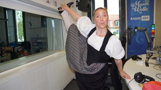 Eine junge Trachtenfrauen zieht mit ihrer rechten Hand ihr rechtes Bein senkrecht in die Höhe.