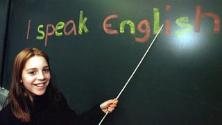 Schülerin zeigt mit Stock auf eine Wandtafel, wo geschrieben steht "I speak English"